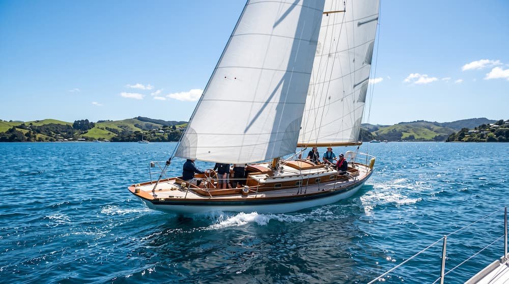 Classic sloop-type sailboat sailing at full sail across a sunny bay.