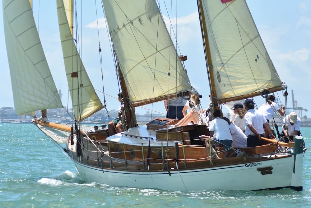 A two-masted wooden sailboat is sailing in turquoise water under a partly cloudy sky. The vessel has cream-colored sails and a crew of several people in white clothing working on the deck.