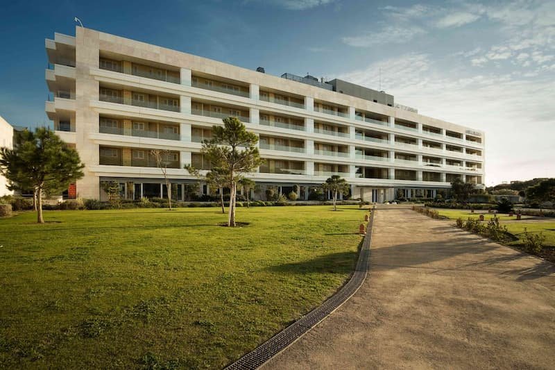 Fachada exterior del moderno edificio de Las Suites en Puerto Sherry, con múltiples plantas, balcones y un jardín verde.